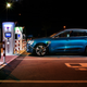 A photo of a teal-colored Audi e-tron electric car charging at a station at night.